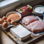 Flat lay of affordable high-protein meal prep ingredients including eggs, canned tuna, dried lentils, frozen chicken thighs, Greek yogurt, and tofu on a wooden cutting board