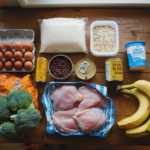 Budget grocery haul flat lay: eggs, rice, oats, canned beans and tuna, frozen broccoli, chicken thighs, bananas, and Greek yogurt on a wooden table